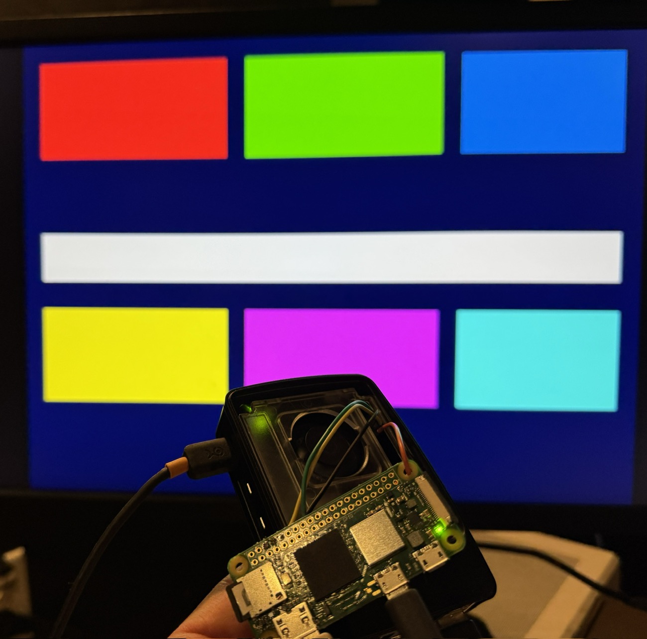 A photograph of a monitor showing a colour test pattern: red, green and blue rectangles across the top, a white bar across the middle, and yellow, magenta and cyan rectangles below, all on a dark blue background. A Raspberry Pi Zero 2 W is held up in front of the screen, wired over USB.