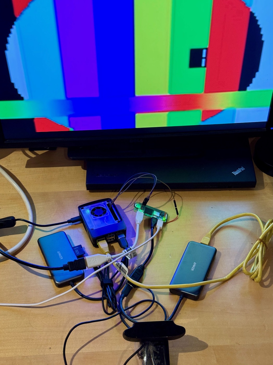 A desk with a Dell monitor showing a decoded video frame — a blocky circle over vertical colour bars with a rainbow gradient band across the middle. Below it a Raspberry Pi 5 in a fan case glows blue beside a bare Pi Zero 2 W with jumper wires attached, two USB hubs, a yellow Ethernet cable, and a webcam on a stand pointed at the screen.