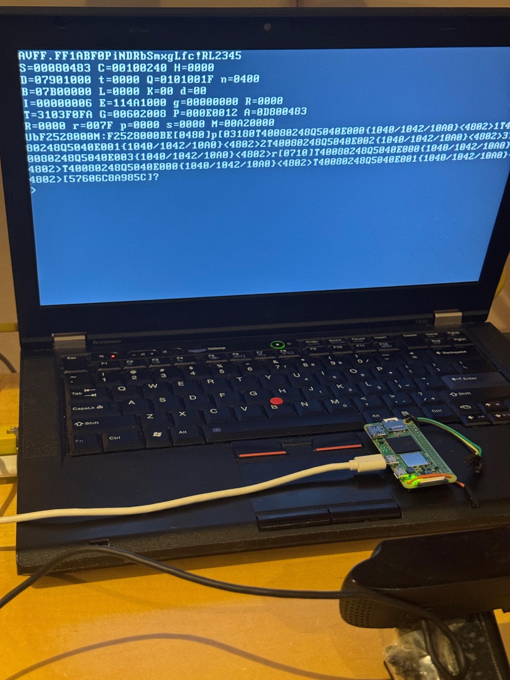 A photograph of a Lenovo ThinkPad T420 with its screen full of blue text: a register dump in hexadecimal and a bare prompt. A Raspberry Pi Zero 2 W board sits on the keyboard, connected by a USB cable, with jumper wires running off it.